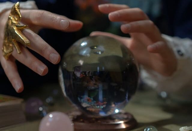 Foto de uma bola de cristal sobre a mesa com uma mulher colocando suas mãoes em cima e ao lado dessa bola. A foto ilustra o Artigo Qual a diferença entre Misticismo e Esoterismo? do site Ori Mystyco.