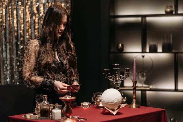 Foto de uma mulher tatuada em pé em frente a uma mesa cheia de elementos esotéricos e místicos. A foto ilustra o Artigo Qual a diferença entre Misticismo e Esoterismo? do site Ori Mystyco.