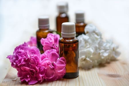 Foto de 4 vidros de óleos essencial. Os vidros têm cor âmbar e tampa branca e estão sobre uma mesa de madeira clara. Na mesa também estão flores brancas e cor de rosa. A foto ilustra o artigo "O que é aromaterapia e quais seus benefícios?" do site Ori Mystyco.