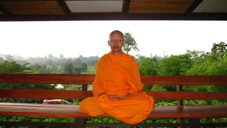 Foto de um monge budista com roupa laranja, sentado meditando em uma estrutura de madeira. Ao fundo se vê árvores. A foto ilustra o artigo "Meditação como exercício saudável e espiritual" do site Ori Mystyco.