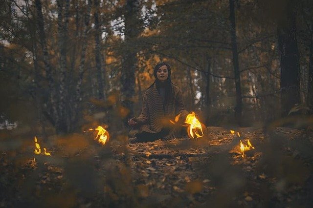 Foto de uma mulher com malha de tricot bege, sentada, meditando no meio da Floresta. na frete dela há 2 pequenas fogueiras de cada lado. A foto ilustra o artigo "O Neoxamanismo e o auge do xamanismo psicodélico" do site Ori Mystyco.