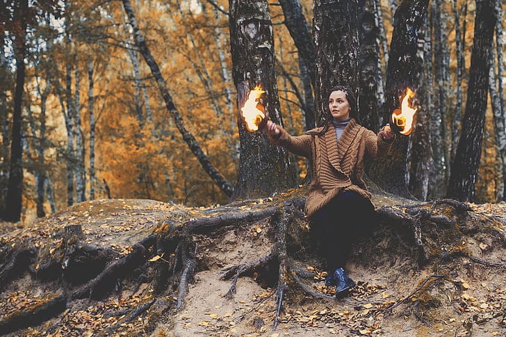 Foto d euma mulher com roupa bege, sentada nas raízes de uma árvore na floresta, com fogo nas mãos. A foto ilustra o artigo "O que é Xamanismo?" do site Ori Mystyco.