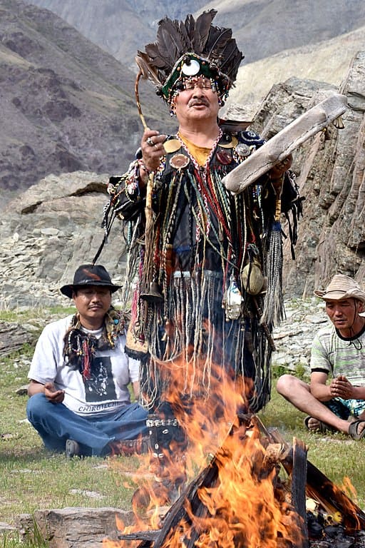 Foto de um xamã sul americano fazendo magia na frente de uma fogueira. A fogueira está em um pasto e atrás dele há um indígena sulamericano sentado e pedras atrás do mesmo. A foto ilustra o artigo "O que é Xamanismo?" do site Ori Mystyco.