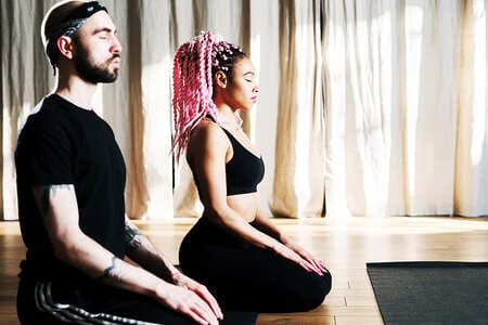 Foto de um casal com roupa preta meditando juntos em uma sala de aula. A foto ilustra este artigo do site Ori Mystyco.