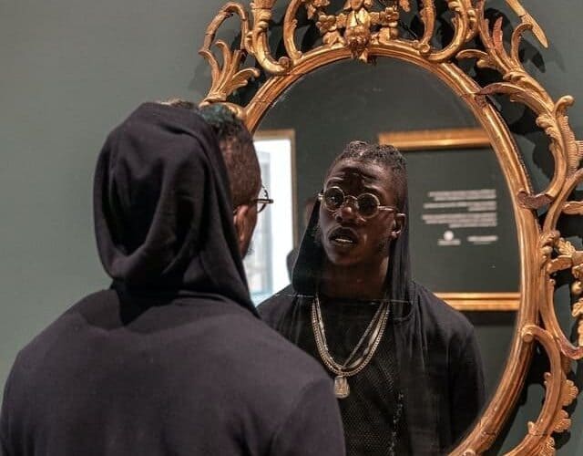 Foto de um homem preto com um moletom preto com capuz. O capuz está na cabeça dele. Ele olha para um espelho oval com moldura dourada. A foto ilustra o artigo A coragem de ser você mesmo do site Ori Mystyco.