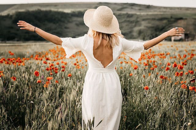 Foto de uma mulher de vestido e chapéus brancos em um campo de flores vermelhas. A mulher está com os braços abertos naturalmente A foto ilustra o artigo Comportamento natural - O ser humano perdeu o seu do site Ori Mystyco.