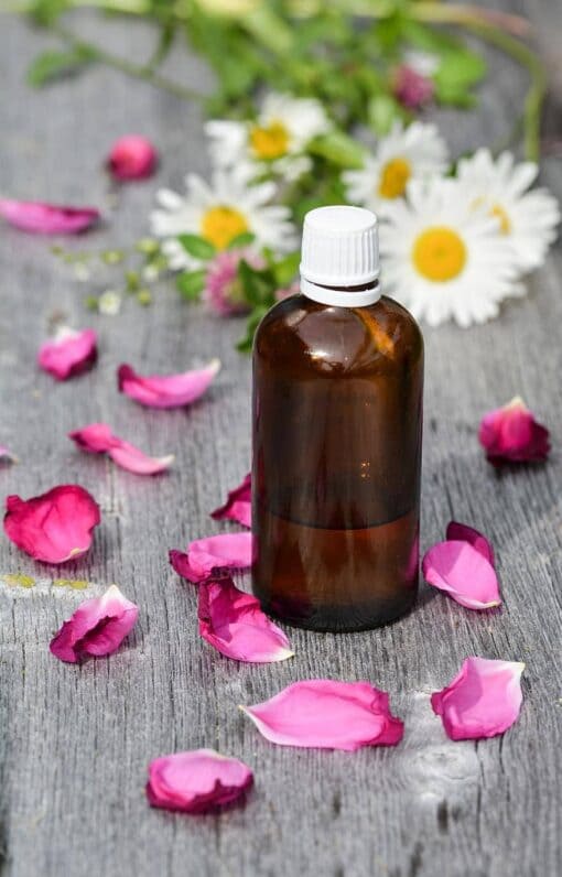 Foto com uma garrafa de vidro âmbar e tampa branca. A garrafa está em cima de uma mesa de madeira acinzentada, onde pode-se ver pétalas de rosa cor-de-rosa e margaridas. A foto ilustra o artigo Garrafa de bruxa de mel: para que serve e como fazer? do site Ori Mystyco.