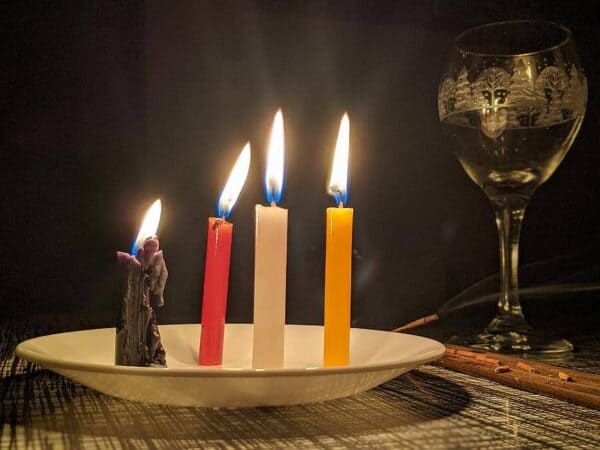 Foto com uma vela palito preto, uma vermelha, uma branca e uma amarela. As velas estão acesas e em cima de uma mesa. Ao lado há uma taça de cristal com bebida. Todos os elemtnos estão em cima de uma mesa. A foto é usada para ilustrar o artigo Magia das Velas. Como praticá-la e quando? do site Ori Mystyco.