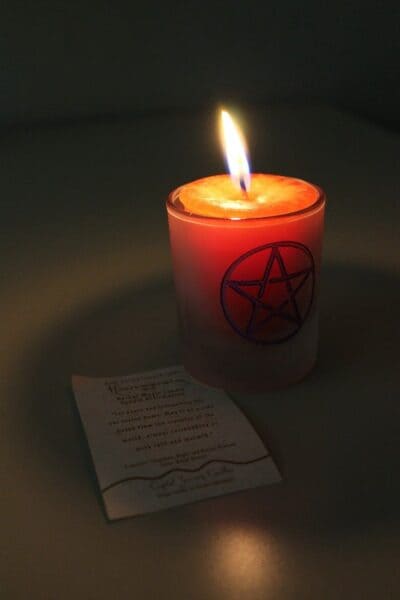 Fptp de uma vela em copo de vidro. No copo de vidro há o desenho de um pentagrama. Em cima da mesa há ainda um papel. A foto é usada para ilustrar o artigo Magia das Velas. Como praticá-la e quando? do site Ori Mystyco.