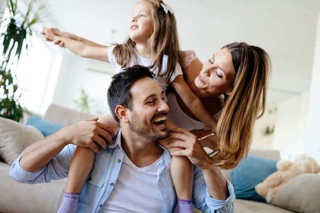 Foto de pai sentado no chão, carregando a filha nos ombros. Atrás da menina está a mãe. A foto serve para ilustrar este artigo do site Ori Mystyco