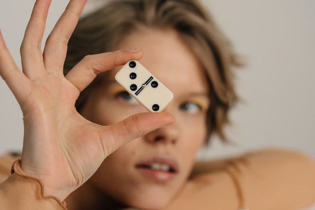 Imagem de mulher loira segurando uma peça de dominó na mão em frente ao rosto. A foto serve para ilustrar a Dominomancia online grátis ou Leitura de Dominós Online gratuita do site Ori Mystyco.