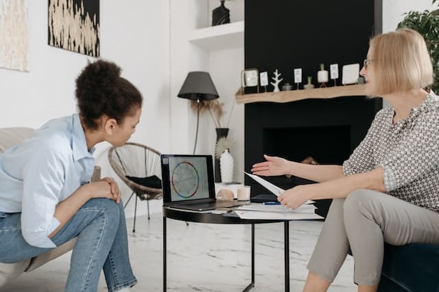 Foto de uma mulher sentada com um mapa astral na mão e o mesmo mapa está sendo projetado na tela de um notebook que está em cima de uma mesinha de centro. A Astróloga está apresentando o mapa astral para outra mulher.. Essa foto serve para ilustrar o artigo Astrologia: Entenda o Papel de um Astrólogo do site Ori Mystyco.