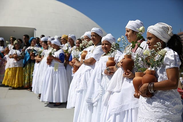 Foto de mulheres pretas com roupa e lenço de renda branca. As mulheres carregam quartinhas de barro com flores dentro. A foto ilustra o artigo O que é Candomblé do site Ori Mystyco.